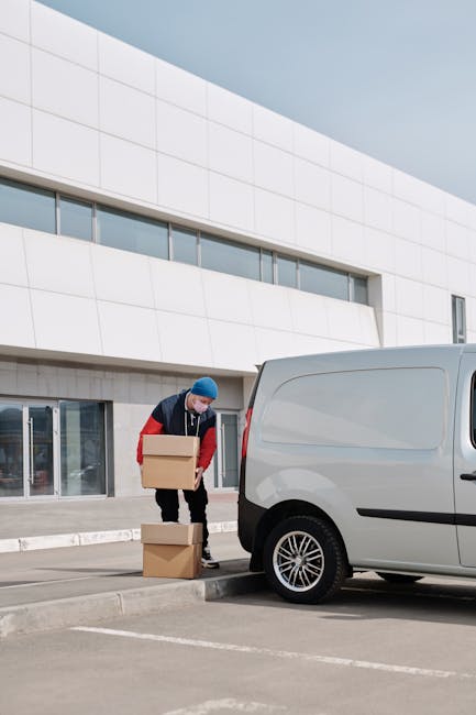 A man dressed in a dark jacket, red sleeves, and a blue beanie is outdoors in front of a modern, white building with horizontal glass windows. He is bending down, carefully placing a small cardboard box onto the ground near a white cargo van parked in a designated loading area. The van's side door is closed, and the vehicle's wheel and part of its rear are visible. Several additional cardboard boxes are positioned on the pavement nearby, indicating a home relocation or moving process. The background shows part of the building's exterior, which is composed of large, smooth white panels, with natural daylight illuminating the scene. The setting reflects a professional removals environment, with [COMPANY_NAME] potentially involved in the packing and furniture transport for the move, as per the context of house removals and logistics.