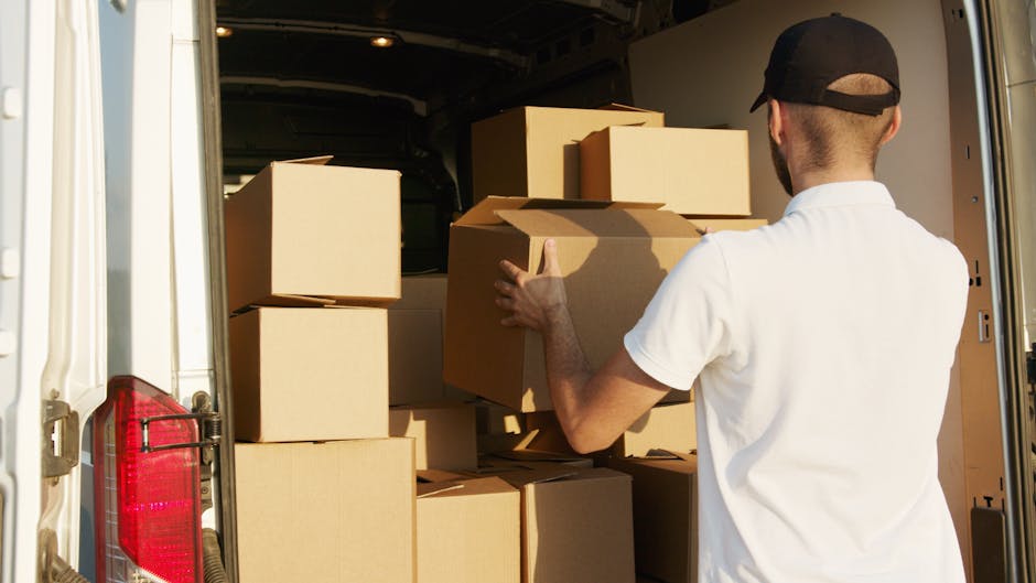 A man wearing a white polo shirt and a black cap is inside the back of a large moving van, stacking and organizing multiple cardboard boxes of various sizes. The boxes are sealed with packing tape and are arranged on the van's floor and shelves, with some placed on top of others to maximize space. The interior of the van is illuminated by natural light through the open rear doors, revealing a smooth, grey lining. The man appears to be preparing the boxes for transport during a home relocation, with the van positioned on the pavement adjacent to a property. Clapham Movers, a professional removals company, manages the packing, loading, and transportation process as part of their house removals service, supporting efficient furniture transport and moving logistics.