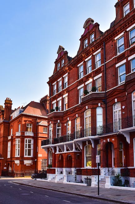 A street view of a multi-storey red-brick Victorian terrace in Clapham, showing the building's ornate architectural details and large sash windows. The property features a small front yard with black iron railings and a white porch canopy supported by decorative brackets. The pavement in front has a parking sign, and the street appears quiet with no visible pedestrians or vehicle activity at the moment. Behind the main building, neighboring similar period homes are visible under a clear blue sky. In the context of house removals, Clapham Movers may use such properties for home relocation or furniture transport services, with trucks or moving equipment typically positioned nearby for loading or unloading. The image captures the residential character relevant to relocation processes within the area, highlighting the typical architecture and environment associated with period homes and flats in Clapham.