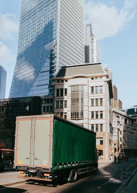 A large green delivery truck with a closed cargo area is parked on a city street, positioned near a sidewalk with a few pedestrians. The truck is situated in front of a modern glass office building wi