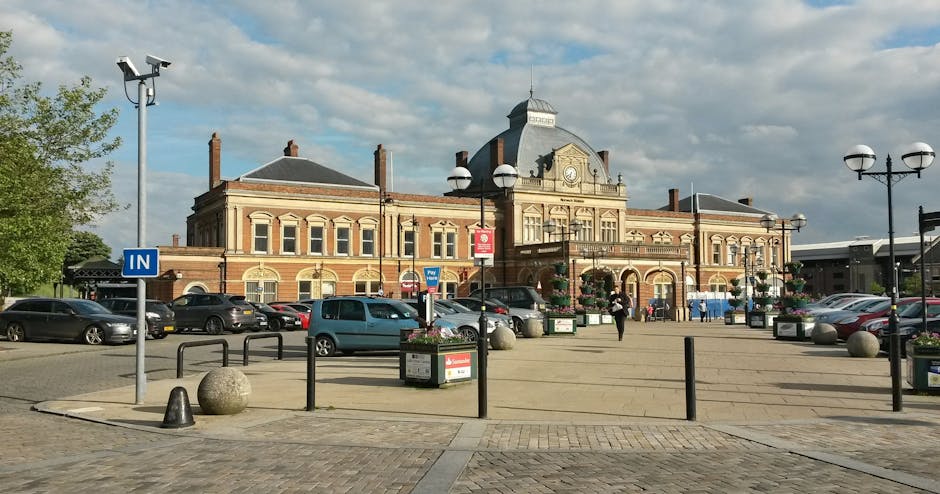 A spacious parking area in front of a historic brick building with a central tower and arched windows, serving as a station or public facility near Clapham Junction. The parking lot contains multiple parked cars of various sizes, including sedans and small vans, arranged along the designated parking spaces. Several large spherical stone bollards and waist-high black metal barriers are positioned along the pavement to direct traffic flow and pedestrian movement. Flower arrangements in rectangular planters are placed on the pavement near lampposts, which are equipped with dual-globe streetlights on black metal poles, providing illumination during evening hours. A blue sign indicating 'IN' directs vehicles entering the parking area, and a metal CCTV camera is mounted on a tall pole in the upper left corner, monitoring the area. The environment appears well-maintained with clean surfaces, and a few pedestrians are visible walking across the open space, possibly engaged in home relocation or moving logistics activities, as associated with Clapham Movers' removals services.
